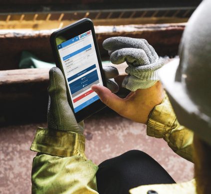 Individual wearing a hard hat and gloves, engaged with a smartphone to access Drive NDT software for work tasks.